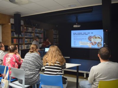 Grupa osób w bibliotece podczas spotkania Kawiarenki Językowej obserwuje prezentację multimedialną o Turcji. Na ekranie widoczny jest slajd dotyczący tradycyjnego jedzenia ulicznego pt. „Tasting authentic street food and local delicacies across Turkey” ze zdjęciami potraw. Prowadzące czuwają przy laptopie z boku sali