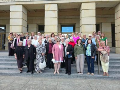 Duża grupa uśmiechniętych kobiet, głównie seniorek, stoi pozując do wspólnego zdjęcia na szerokich, kamiennych schodach przed monumentalnym budynkiem z jasnymi, klasycznymi kolumnami. Kobiety są ubrane w eleganckie, letnie stroje casualowe w różnych kolorach i wzorach