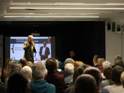 Jacek Fedorowicz przemawia do mikrofonu na scenie w bibliotece. Za nim widoczny jest ekran projekcyjny z plakatem wydarzenia oraz siedzący prowadzący. Na pierwszym planie widać tyły głów zgromadzonej publiczności