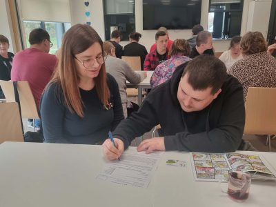 Szatynka w okularach wypełnia formularz przy stole obok starszej kobiety, obie uważnie patrzą na dokument. W tle widoczny wózek inwalidzki oraz otwarte drzwi do sali