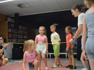 Dzieci i opiekunowie biorą udział w zabawie ruchowej w sali bibliotecznej. Dwie dorosłe osoby trzymają napięty żółty sznur, pod którym przechodzą dzieci stojące w kolejce na dywanie w biało-czerwone pasy. W tle widać regały z książkami