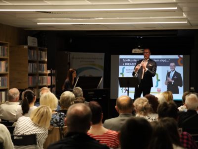 Mężczyzna w garniturze gra na klarnecie na scenie w bibliotece. Obok, przy pianinie cyfrowym CASIO, siedzi kobieta. W tle widać publiczność oraz ekran z napisem: Spotkanie z cyklu „Jestem stąd…” z Łukaszem Motyką
