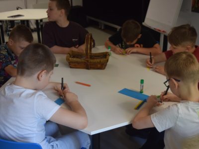Zbliżenie na grupę sześciu chłopców siedzących wokół białego stołu i zaangażowanych w rysowanie. Na blacie leżą kredki, kleje w sztyfcie oraz paski kolorowego papieru. Na środku stołu stoi podłużny, wiklinowy koszyczek z przyborami plastycznymi