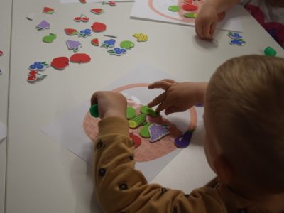 Zbliżenie na rączki dziecka przy białym stoliku, które przykleja kolorowe, piankowe naklejki w kształcie owoców – truskawek, winogron i jabłek – na kartkę z rysunkiem koszyka