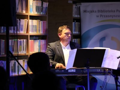 Mężczyzna w garniturze gra na instrumentach klawiszowych Korg umieszczonych na scenie. Obok niego stoi regał pełen książek oraz roll-up Miejskiej Biblioteki Publicznej w Przasnyszu