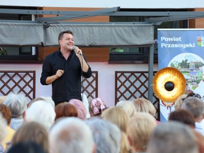 Robert Kudelski śpiewa na scenie pod zadaszeniem z materiału. Artysta uśmiecha się, trzymając mikrofon w prawej ręce i delikatnie gestykulując drugą dłonią. Za jego plecami znajduje się baner promocyjny Powiatu Przasnyskiego ze zdjęciami zabytków i krajobrazów. Pierwszy plan zajmują głowy widzów wsłuchanych w występ
