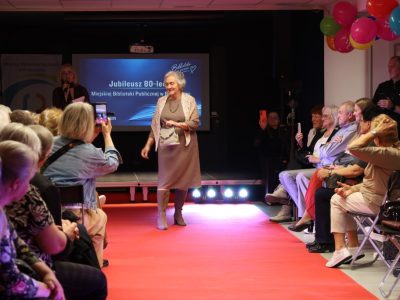 Kobieta w klasycznej beżowej sukience i jasnym, dzianinowym szalu nakrytym na ramiona prezentuje małą, wzorzystą torebkę do ręki. Widzowie siedzący po bokach nagrywają pokaz telefonami