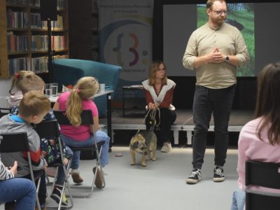 Mężczyzna stojący na środku sali przemawia do dzieci, podczas gdy Marta Maj siedzi na podwyższeniu i trzyma na smyczy niewielkiego, kundelka o imieniu Precel