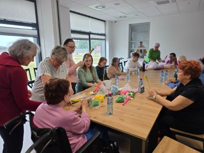 Grupa dorosłych osób siedzi wokół stołu, gdzie tworzą palmy wielkanocne. Na stole znajdują się materiały potrzebne do stworzenia świątecznej ozdoby.