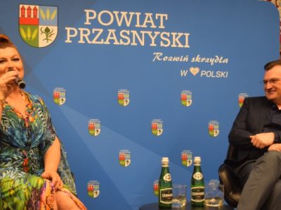 Katarzyna Skrzynecka mówi do mikrofonu, siedząc na fotelu obok prowadzącego w ciemnej marynarce, który z uśmiechem jej słucha. Na małym stoliku przed nimi stoją butelki z wodą