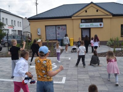 Grupa dzieci bawi się na wybrukowanym placu przed parterowym, żółtym budynkiem z szyldem „DOM HARCERZA”. Na nawierzchni placu widoczne jest namalowane białą farbą oznakowanie miejsca dla osób z niepełnosprawnościami