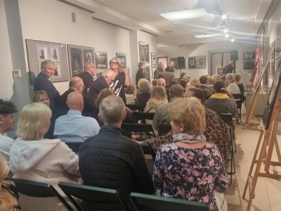 Widok od tyłu na widownię wypełniającą salę wystawową podczas wernisażu. Po prawej stronie stoją drewniane sztalugi, a na ścianach wiszą oprawione fotografie i obrazy