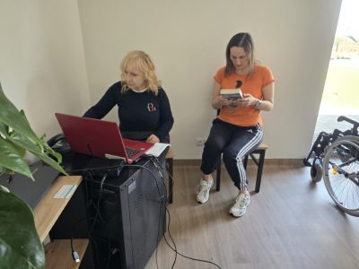 Wnętrze pomieszczenia, w którym dwie kobiety zajmują się pracą z literaturą. Jedna siedzi przy biurku z czerwonym laptopem, druga siedzi obok na krześle i czyta książkę. Po prawej stronie widoczny jest fragment wózka inwalidzkiego, a po lewej zielone liście rośliny doniczkowej