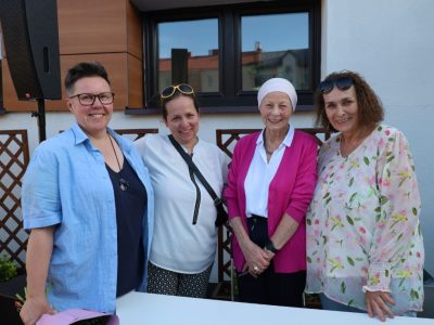 Cztery uśmiechnięte kobiety pozują do zdjęcia za białym stołem na świeżym powietrzu. Druga od prawej to aktorka Anna Nehrebecka, ubrana w fuksjowy kardigan, białą bluzkę i jasny turban na głowie. Pozostałe uczestniczki stoją obok niej w letnich strojach. W tle widać fragment budynku oraz drewniane kratki ogrodowe