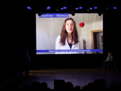 Widok na scenę, na której duży ekran wyświetla nagranie wideo z młodą kobietą składającą życzenia („Josefine - Niemcy”) z umieszczonymi na dole napisami. Po prawej stronie sceny siedzi dyrektor biblioteki