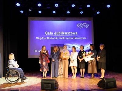 Siedem kobiet tworzących delegację stoi w rządku na scenie, trzymając bukiety kwiatów i upominki. Pierwsza z nich przemawia do mikrofonu, zwracając się do siedzącej po lewej stronie dyrektor biblioteki
