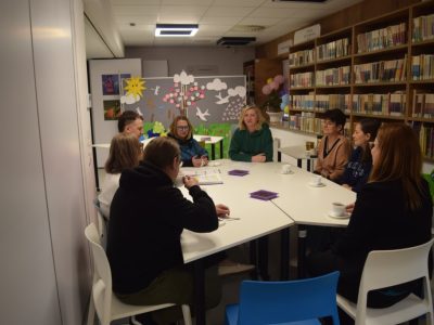 Widok na salę biblioteczną, w której osiem osób uczestniczy w spotkaniu przy wspólnym stole. Część uczestników pije napoje z białych filiżanek, na stole leżą też drobne poczęstunki w fioletowych pojemnikach. Ściany pomieszczenia zdobią półki z książkami oraz dziecięce rysunki z napisem „Wiosna”