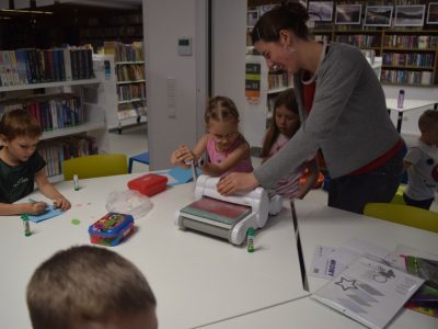 Zbliżenie na wolontariuszkę pomagającą dziewczynce w obsłudze białej maszynki do scrapbookingu. Dziewczynka kręci korbką, aby wyciąć kształty w papierze. Obok na stole inne dziecko przykleja elementy do swojej kartki. W tle znajdują się biblioteczne regały wypełnione książkami