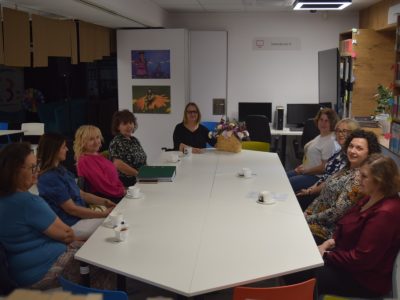 Uczestniczki spotkania siedzą przy białym stole w sali bibliotecznej, patrząc w stronę obiektywu i uśmiechając się. Na środku stołu, obok filiżanek, stoi wiklinowy kosz z fioletowo-białymi kwiatami. W tle widoczne są regały z książkami oraz komputery