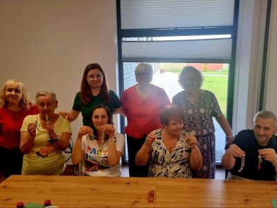 Osiem osób – pięć stojących z tyłu i trzy siedzące z przodu przy długim drewnianym stole – pozuje do zdjęcia w jasnym pomieszczeniu. Uczestnicy uśmiechają się i trzymają w dłoniach małe, ręcznie robione breloczki ze sznurka (przypominające postacie lub plecionki)