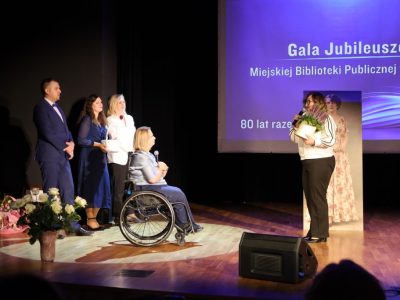 Kobieta w jasnej bluzce przemawia do mikrofonu, trzymając w dłoniach białą doniczkę z rozkwitającymi kwiatami, zwrócona w stronę dyrektor biblioteki oraz towarzyszących jej prowadzących