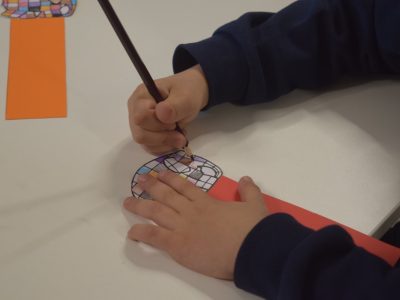 Zbliżenie na dłonie dziecka w granatowej bluzie, siedzącego przy białym stole. Dziecko starannie koloruje ołówkiem precyzyjny, geometryczny wzór na papierowym elemencie w kształcie koła, który jest przymocowany do czerwonego paska papieru