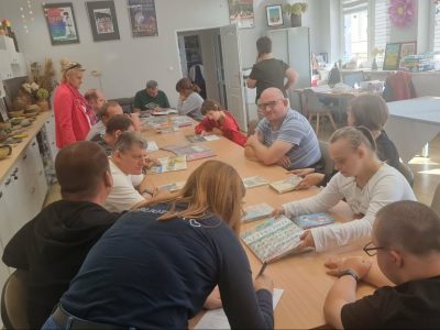 Szerokie ujęcie z góry na grupę kilkunastu osób siedzących przy długim, drewnianym stole w jasnej świetlicy. Uczestnicy, w tym osoby dorosłe i młodzież, przeglądają rozłożone na stole książki i ilustracje. Na pierwszym planie widać kobietę w ciemnym ubraniu. W tle widać białe szafki z wystawą rękodzieła (ceramika, suche bukiety) oraz plakaty na ścianach. Panuje swobodna atmosfera skupienia