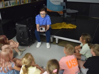 Kobieta w okularach i niebieskiej koszulce w paski siedzi na podwyższeniu, pokazując mały, retro projektor grupie dzieci siedzących przed nią w półkolu na podłodze