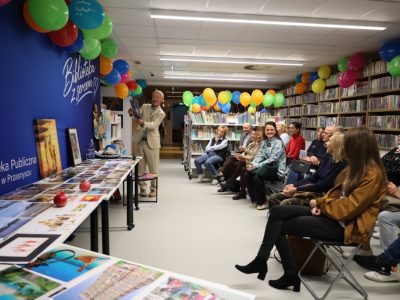Szerokie ujęcie sali bibliotecznej wypełnionej słuchaczami. Andrzej Ciuba stoi przy długim stole wyłożonym zdjęciami i podnosi jedno z nich w górę, prezentując je z uśmiechem uśmiechniętym widzom