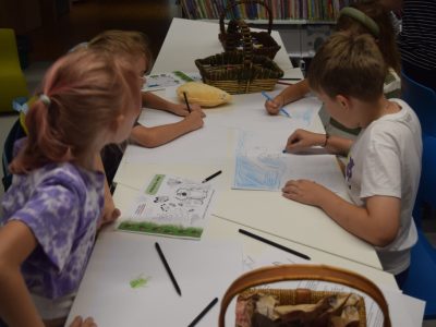 Grupa dzieci siedzi przy podłużnym, białym stole i rysuje kredkami na białych kartkach papieru. Na stole leżą materiały edukacyjne oraz wiklinowe koszyki z przyborami plastycznymi
