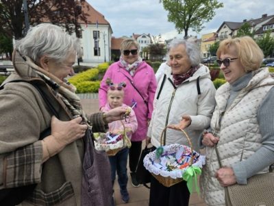 Grupa kobiet i dziewczynka rozmawiają na miejskim placu. Jedna z kobiet trzyma koszyk z upominkami, z którego inna osoba wybiera podarunek. W tle widoczna jest architektura rynku oraz drzewa. Uczestniczki spotkania są uśmiechnięte i ubrane w wiosenne kurtki oraz kamizelki