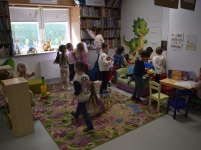 Wnętrze biblioteki dla dzieci. Dzieci bawią się w strefie przeznaczonej do zabawy: część dzieci siedzi na kolorowym dywanie, inne korzystają z klocków i zabawek, a jeszcze inne siedzą przy małym stoliku z książkami. Przy regale stoi dorosła osoba, porządkująca książki. Na ścianie widoczna jest duża grafika smoka