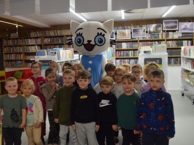 Grupa kilkanaściorga przedszkolaków pozuje do pamiątkowego zdjęcia w bibliotece. W samym centrum, za dziećmi, stoi duża maskotka białego kota w niebieskiej sukience (Kicia Kocia). W tle widoczne są regały wypełnione kolorowymi książkami dla dzieci