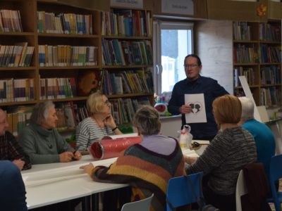 Spotkanie warsztatowe w bibliotece. Mężczyzna w ciemnej koszuli stoi na tle regałów i prezentuje siedzącym przy stole uczestnikom kartkę z szablonem graficznym – czarnymi sylwetkami kobiety i mężczyzny wewnątrz serca. Zebrani patrzą w jego kierunku i słuchają instrukcji