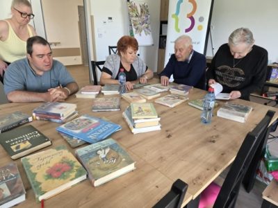 Uczestnicy zajęć bibliotecznych skupieni wokół stołu z książkami. Na blacie leżą m.in. „Podróże Guliwera”, „Tajemniczy ogród” oraz „Słownik”. Osoby przy stole rozmawiają i wspólnie przeglądają literaturę. W tle widoczny biały roll-up biblioteki oraz tablica z kolorową pracą plastyczną