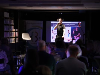 Damian Kowalski podczas koncertu w przyciemnionej sali bibliotecznej. Wokalista dynamicznie śpiewa na scenie, trzymając mikrofon. W tle widoczny jest ekran z plakatem koncertu, a przed sceną siedzą widzowie, z których jeden nagrywa występ telefonem