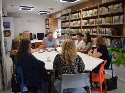 Uczestnicy spotkania w bibliotece uwiecznieni z innej perspektywy. Mężczyzna w błękitnej koszulce opiera się o stół i mówi, wykonując gest ręką, podczas gdy reszta grupy słucha go w skupieniu. Po prawej stronie, na ścianie pod drewnianymi regałami pełnymi książek, zamontowane są kolorowe, ścienne zabawki manipulacyjne dla dzieci (m.in. labirynt i ksylofon)