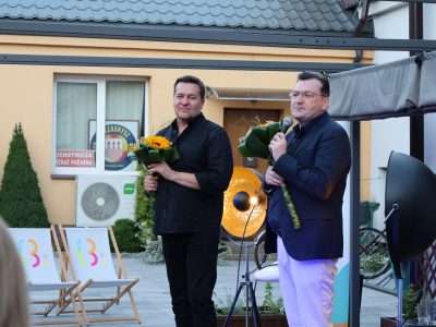 Robert Kudelski oraz prowadzący spotkanie stoją na scenie po zakończeniu występu, trzymając w rękach bukiety żółtych słoneczników. Kudelski, w czarnej koszuli, uśmiecha się łagodnie w stronę widowni. Prowadzący w ciemnogranatowej marynarce, jasnych spodniach i okularach przemawia do mikrofonu. W tle widoczny jest fragment budynku z godłem i napisem „Ochotnicza Straż Pożarna” oraz leżaki biblioteczne