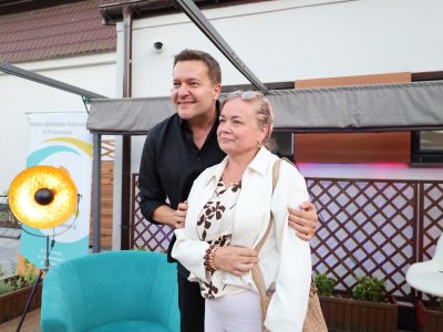 Robert Kudelski pozuje do pamiątkowej fotografii z kobietą na scenie po wydarzeniu. Artysta w czarnej koszuli obejmuje ramieniem uśmiechniętą uczestniczkę, która ma na sobie białą marynarkę, bluzkę we wzory i jasne spodnie. W tle widać dekoracyjną, złotą lampę oraz drewniane kratki na ścianie budynku