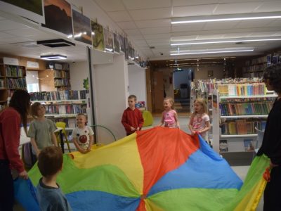 Grupa dzieci i bibliotekarka trzymają za brzegi dużą, kolorową chustę animacyjną w przestrzeni bibliotecznej. Wszyscy są uśmiechnięci i przygotowani do wspólnej zabawy ruchowej