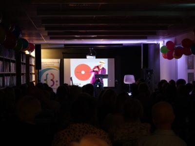 Szerokie ujęcie zaciemnionej sali z widownią ukazaną w sylwetkach. Na oświetlonej scenie w głębi stoi Stanisław Górka w słomkowym kapeluszu i marynarce, śpiewając na tle ekranu z grafiką projektu „DROGA”