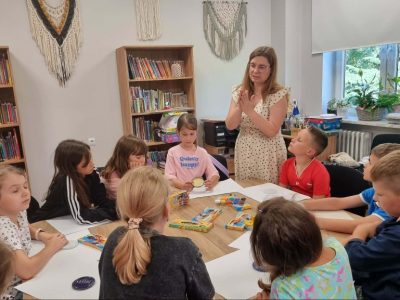 Grupa dzieci siedzi przy drewnianym stole w jasnej sali bibliotecznej i słucha stojącej przed nimi instruktorki w okularach oraz jasnej sukience w groszki, która gestykuluje dłońmi. Na stole leżą opakowania z plasteliną i białe kartki papieru