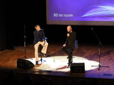 Dwaj mężczyźni siedzą na wysokich stołkach barowych w plamie światła na scenie i prowadzą rozmowę do mikrofonów. W tle na ekranie widnieje napis „80 lat razem”