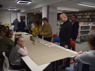 Uczestnicy warsztatów siedzą wzdłuż długiego stołu pokrytego szarym papierem ochronnym. Dwie kobiety stoją na środku, rozstawiając butelki z kolorowymi farbami plakatowymi i papierowe talerzyki. Pozostałe osoby obserwują przygotowania. W tle widać jasną ścianę z dekoracją przedstawiającą napis „JESIEŃ” i słoneczniki