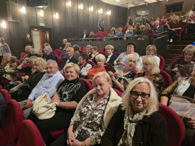 Uczestnicy wyjazdu siedzą w czerwonych fotelach na widowni teatralnej i uśmiechają się w stronę aparatu. W tle widać zapełniające się rzędy widowni