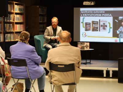 Widok z perspektywy widowni na Krzysztofa Piersę siedzącego w fotelu z filiżanką. Za nim znajduje się ekran z prezentacją z tytułem spotkania autorskiego i okładkami książek