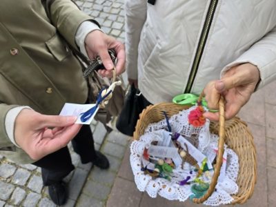Zbliżenie na dłonie dwóch osób na chodniku. Jedna osoba trzyma mały upominek z dołączoną karteczką i niebieską wstążką. Druga osoba trzyma wiklinowy koszyk wyścielony koronkową serwetką, wypełniony podobnymi upominkami i bilecikami